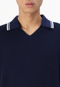 Polo din tricot de culoare bleu marin, cu decolteu în V, având o guler cu dungi albe contrastante și un material texturat. Vedere detaliată a jumătății superioare.