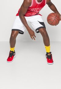 Punainen koripallopaita, jossa on teksti "CHICAGO", valkoiset shortsit mustilla yksityiskohdilla, keltaiset sukat ja punamustat lenkkarit. Pitelee koripalloa.