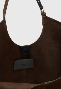 Ruskea mokkanahkainen olkalaukku, jossa on musta nahkahihna ja sisämerkki. Laukussa on magneettinen solki ja metallinen solkiyksityiskohta.