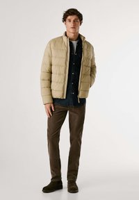Chaqueta acolchada beige con cierre frontal, cuello alzado y diseño acolchado, combinada con una camisa de mezclilla oscura y pantalones marrones, completando con botas verdes.