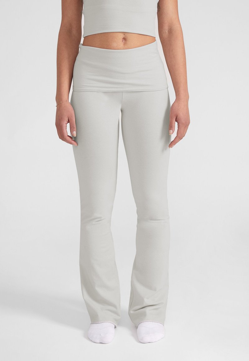 Donna che indossa leggings grigio chiaro a vita alta e calzini bianchi, in piedi su uno sfondo chiaro e uniforme.