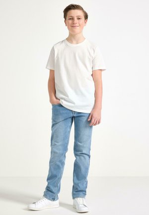 Ragazzo adolescente sorride con una mano in tasca, indossando una maglietta bianca semplice, jeans azzurri e sneakers bianche, su uno sfondo bianco.