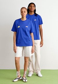 Blauwe katoenen t-shirts met grafische ontwerpen, gecombineerd met witte gestreepte shorts en broeken. Schoeisel heeft zwarte en witte vetersluiting details.