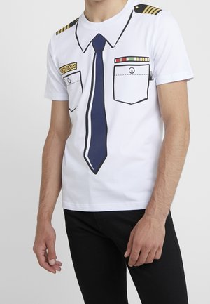 Hombre con camiseta blanca que lleva una corbata azul marino impresa, hombreras de capitán y bolsillos en el pecho con insignias.