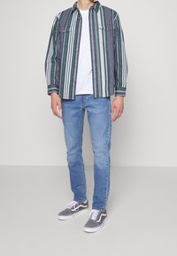 Hombre vestido con vaqueros de mezclilla azul, zapatillas gris y blancas, camiseta blanca y camisa de manga larga a rayas verdes, azules y blancas desabrochada.