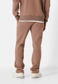 Pantalones de chándal de algodón marrón con un ajuste holgado, que cuentan con un bolsillo trasero y puños acanalados, combinados con zapatillas blancas. Textura suave.