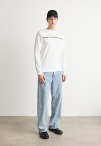 Weißes Langarmhemd mit schwarzer Logo-Streife, hellblaue weite Jeans und schwarze Mütze. Schwarze Schuhe vervollständigen das Outfit, vor einem hellen Hintergrund.