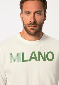 Cremefarbenes Langarm-T-Shirt mit fettem grünen Schriftzug "MILANO" in strukturiertem Design. Weiches Baumwollmaterial, runder Halsausschnitt und lässiger Schnitt.