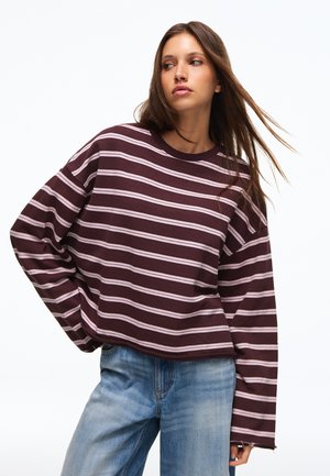 Junge Frau mit langen braunen Haaren, die ein weinrotes und weiß gestreiftes Langarmshirt und hellblaue Jeans trägt, posiert vor einem weißen Hintergrund.