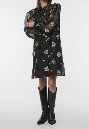 Robe noire transparente avec broderie florale blanche, devant à boutons, manches longues, accompagnée de bottes noires en cuir jusqu'aux genoux.