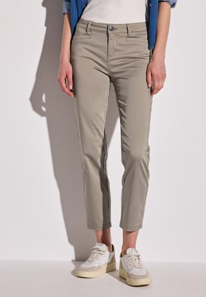 Helle beige Stoffhosen in Slim-Fit, mit fünf Taschen und umgeschlagenen Säumen. Kombiniert mit weißen und grauen Sneaker, vor einem neutralen Hintergrund.