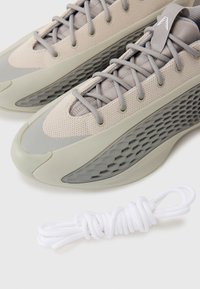 Paire de chaussures de sport beige et grises avec des côtés texturés, des lacets gris et une paire supplémentaire de lacets blancs à côté.