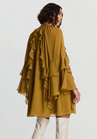 Senfgelbes Kleid mit Rüschärmeln und Rücken-Detail. Leichter, fließender Stoff mit lockerem, knielangem Schnitt. Paar weiße Overknee-Stiefel.