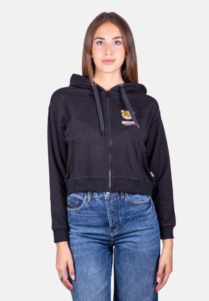 Felpa nera cropped con zip e logo dell'orso sul petto, cappuccio con cordino e polsini a costine. Abbinata a jeans blu a vita alta.