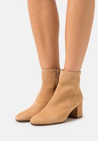 Beige ruskinds ankelstøvler med firkantet tå, en lav blokhæl og en lynlås på siden for nem påføring. Glat tekstur og enkel design.