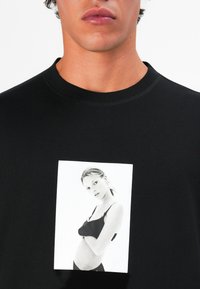Zwarte katoenen t-shirt met een centrale zwart-wit foto print van een vrouw in een zwarte outfit, ronde halslijn en korte mouwen.