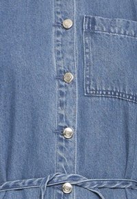 Gros plan d'une chemise en denim bleu avec boutons-pression argentés, poche poitrine et ceinture en tissu nouée autour de la taille.