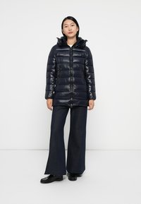 Doudoune marine avec une finition brillante, fermeture éclair sur toute la longueur et une capuche surdimensionnée, associée à un pantalon en denim foncé à jambes larges.