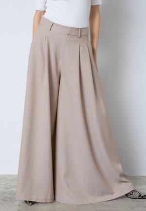 Žena oblečená v béžových širokých nohaviciach s plisovaním, v kombinácii s bielym topom a topánkami so vzorom hadej kože, stojaca vo vnútri na betónovej podlahe.