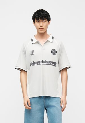 Tânăr cu păr negru purtând un tricou polo alb cu textul „playablanca” și guler cu dungi negre, asortat cu blugi bleu deschis.