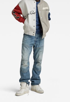 Personne portant un jean bleu clair, des baskets blanches montantes, une chemise bleue et une veste boutonnée à blocs de couleur avec des panneaux rouges, gris et blancs.