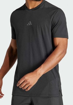 Homme portant un t-shirt de sport noir à manches courtes avec un petit logo Adidas sur la poitrine, debout devant un fond clair uni.