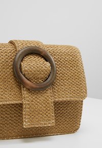 Wovený rovný slamáky v světle hnědé barvě. Obsahuje kulatý dřevěný prsten jako ozdobu a klopu se šitými detaily.