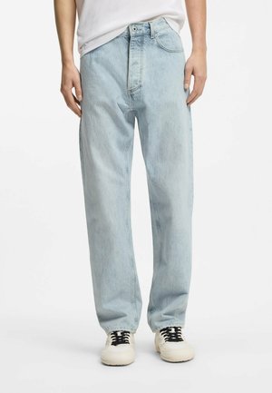 Person wearing light blue loose-fit jeans, white sneakers with black laces, and a partially visible white t-shirt, standing against a white background.