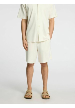 Lys beige kortærmet skjorte med knapper og matchende shorts lavet af blødt stof, suppleret af brune slip-on sandaler. Glat tekstur med minimale detaljer.
