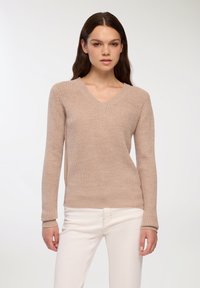 Beżowy sweter z dzianiny z dekoltem w serek i długimi rękawami; fakturowana tkanina i ściągacze u mankietów; noszony z jasnymi spodniami.