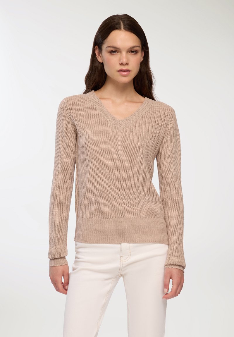 Beżowy sweter z dzianiny z dekoltem w serek i długimi rękawami; fakturowana tkanina i ściągacze u mankietów; noszony z jasnymi spodniami.