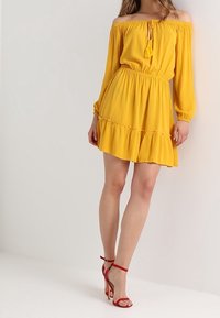 Gelbes schulterfreies Kleid mit langen Ärmeln, elastischem Bund, gerafftem Saum und Quasten-Detail. Kombiniert mit roten Riemchen-Pumps.