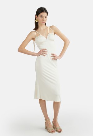 Donna con capelli lisci e scuri indossa un vestito midi bianco a sottoveste e tacchi dorati con cinturini, posando con le mani sui fianchi su uno sfondo semplice.