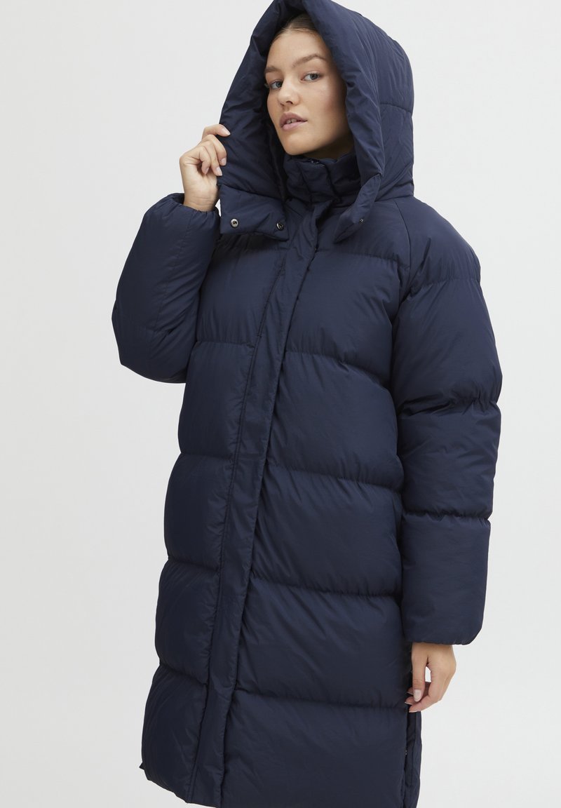 Marineblauwe gewatteerde winterjas met een hoge kraag en grote capuchon. Bevat drukknoopsluitingen en een gebakken textuur voor warmte en comfort.