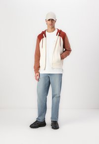 Hettegenser med kremfarget kropp, brune ermer og røde detaljer. Kombinert med blå jeans og svarte joggesko. Strukturet passform, synlige glidelåser.