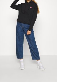Sudadera negra con capucha y bolsillo frontal, jeans de mezclilla azul sueltos y zapatillas deportivas blancas. Sin patrones visibles ni acentos distintivos.