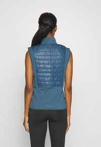 Blå ärmlös väst med en vadderad, quiltad bakpanel. Den släta framdelen kontrasterar med den texturerade baksidan. Enkelt design och ingen synlig hårdvara.