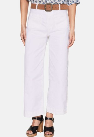Pantalons larges blancs en coton, dotés de deux poches avant et d'une ceinture marron avec une boucle décorative. Portés avec des sandales à plateforme noires.