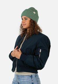 Navy bomber jacket with ribbed cuffs, side pockets, and a front zipper. Paired with a green ribbed beanie. Casual fit and fabric textures visible.