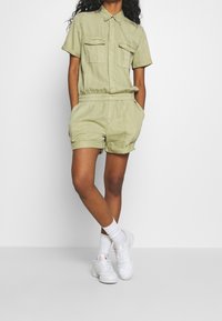 Ljusgrön kort jumpsuit med korta ärmar, två bröstfickor, elastisk midja och uppvikt nederkant. Matchas med vita sneakers och strumpor.