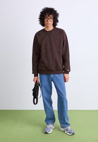 Brown sweatshirt with a textured finish, paired with light blue jeans. Model holds a black bag, stands on green flooring, with a plain background.