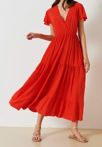Robe maxi rouge avec un décolleté en V et de courtes manches volantes. Elle présente une taille froncée et un ourlet à volants, fabriquée en tissu léger. Sandales montrées.