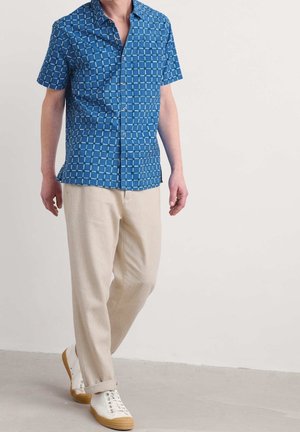 Mann trägt blau gemustertes Kurzarmhemd, beige Hose und weiße Turnschuhe und steht vor einem schlichten hellen Hintergrund.