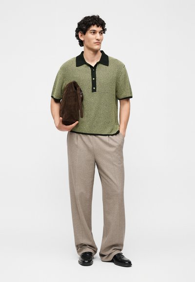 Άνδρας που φοράει πράσινο πλεκτό πόλο, μπεζ παντελόνι με φαρδιά μπατζάκια, μαύρα παπούτσια, κρατώντας καφέ σουέντ τσάντα σε λευκό φόντο.