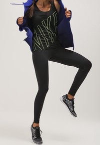 Černý sportovní tank top s neonově zeleným logem, doplněný černými legínami. Modrá bunda, černo-bílé sportovní boty s texturovaným vzorem.