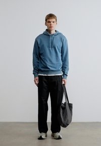 Camisola com capuz azul com cordões, combinada com calças pretas e camisa branca. Saco tote cinzento e ténis atléticos completam o conjunto. Design minimalista.