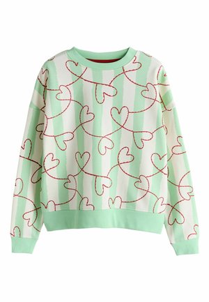 Pull avec des rayures verticales vert clair et crème, motif de lignes en forme de cœur rouge, porté avec un jean bleu clair, main dans la poche.