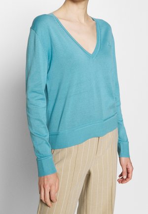 Femme portant un pull bleu clair à col en V et un pantalon beige à fines rayures, vue du cou aux hanches sur un fond uni.