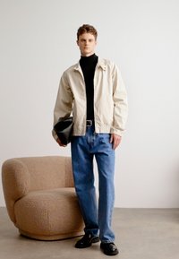 Helle beige Jacke über einem schwarzen Rollkragenpullover, kombiniert mit blauen Jeans und schwarzen Schuhen. Hält eine schwarze Tasche. Weicher beiger Stuhl im Hintergrund.