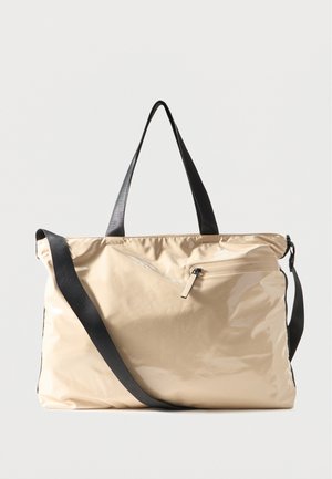 Bēšs spīdīgs liels rokassoma ar melnu plecu siksnu un rokturiem, priekšējā rāvējslēdzēja kabata, attēlota uz vienkārša gaiša fona.